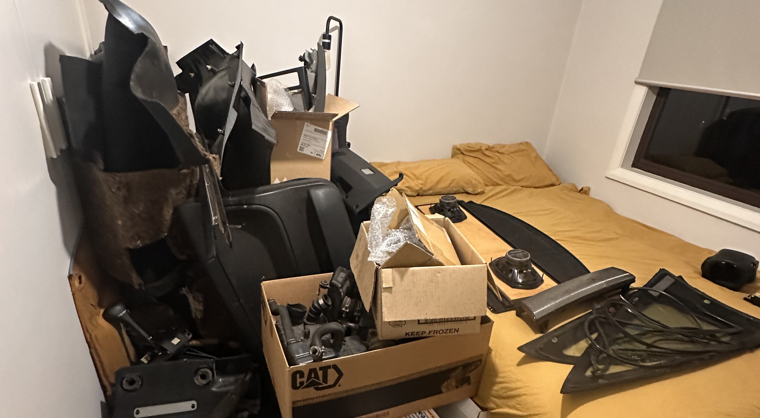 A bedroom with car parts stacked on and around a bed. Cardboard boxes filled with hoses, vents, and components sit on a mustard yellow bedspread, alongside interior trim pieces, taillights, and panels leaning against the wall.